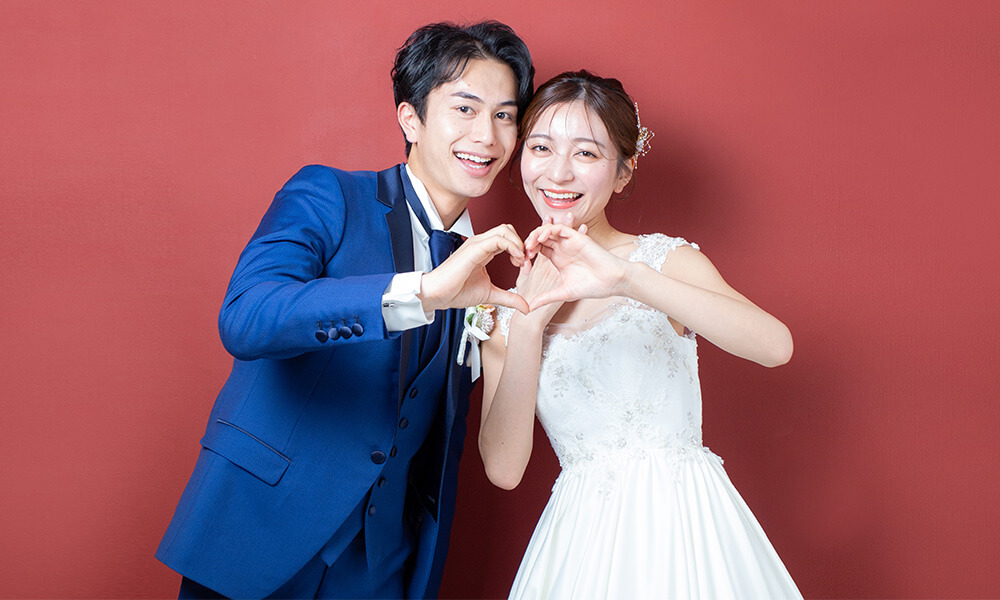 赤い背景の「マイブライド一宮」のスタジオで洋装フォトを撮影する花嫁と新郎のイメージ