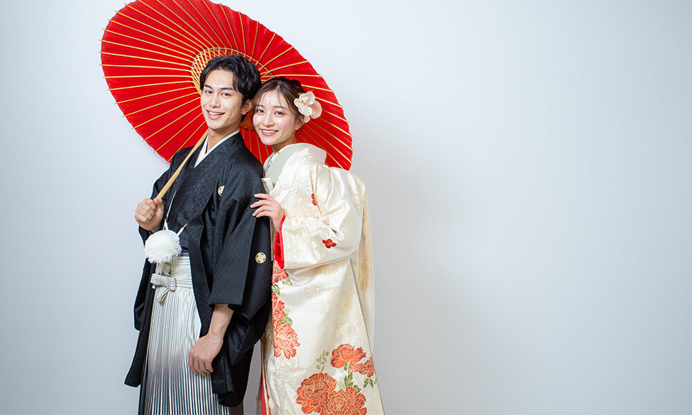 グレーの背景の「マイブライド一宮」のスタジオで赤い和傘を持って和装フォトを撮影する花嫁と新郎のイメージ
