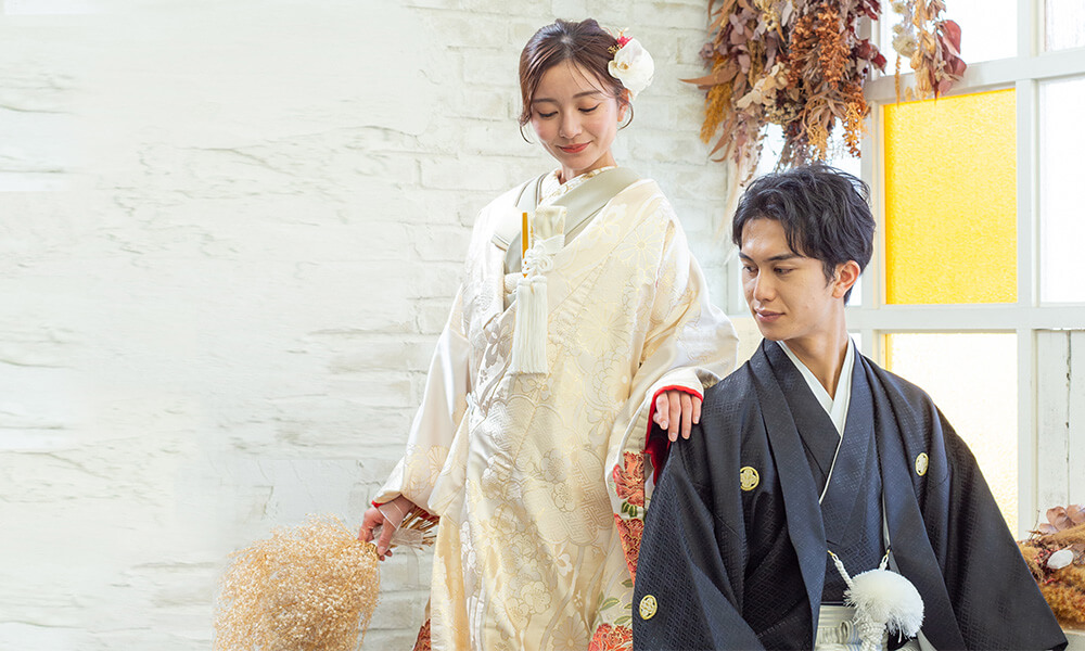 ドライフラワーと白のレンガ背景の「マイブライド一宮」のスタジオで和装フォトを撮影する花嫁と新郎のイメージ