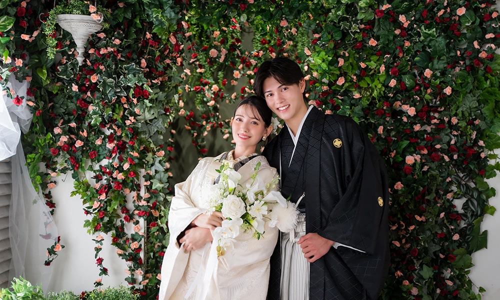 バラの生えた背景の「マイブライド春日井」のスタジオで和装フォトを撮影する花嫁と新郎のイメージ