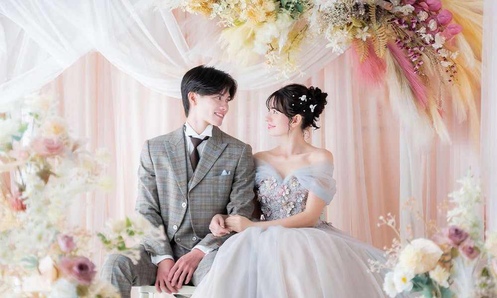 淡いピンクのレースカーテンとドライフラワーなどが配置された「マイブライド春日井」のスタジオで洋装フォトを撮影する花嫁と新郎のイメージ