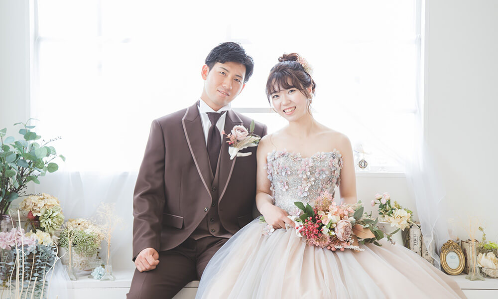 白の窓の前に花や小物が並べてある「マイブライド清洲」のスタジオで座って洋装フォトを撮影する花嫁と新郎のイメージ