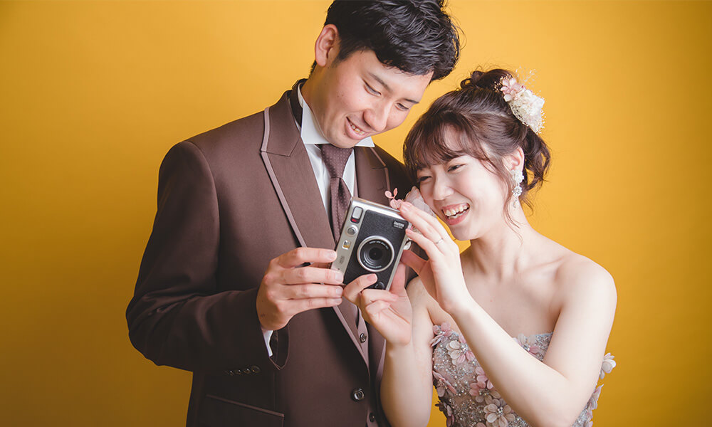 黄色の背景の「マイブライド清洲」のスタジオでレトロカメラを一緒に見ているシーンの洋装フォトを撮影する花嫁と新郎のイメージ