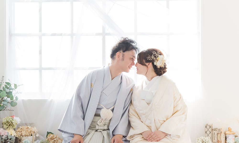 白の窓の前に花や小物が並べてある「マイブライド清洲」のスタジオで座って顔を向かい合わせて和装フォトを撮影する花嫁と新郎のイメージ