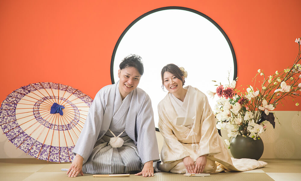赤い壁に生花を配置した和の風情あふれる「マイブライド清洲」のスタジオで正座をして和装フォトを撮影する花嫁と新郎のイメージ