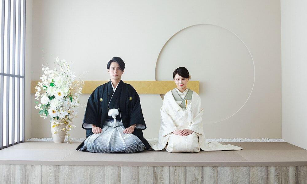 優しい光がさしこむ白い背景の「マイブライド岡崎光ヶ丘店」のスタジオで正座で和装フォトを撮影する花嫁と新郎のイメージ