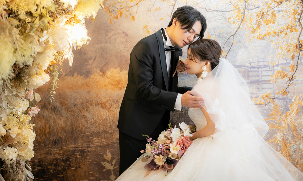 背の高いドライフラワーの配置された「マイブライド岡崎光ヶ丘店」のスタジオで洋装フォトを撮影する花嫁と新郎のイメージ