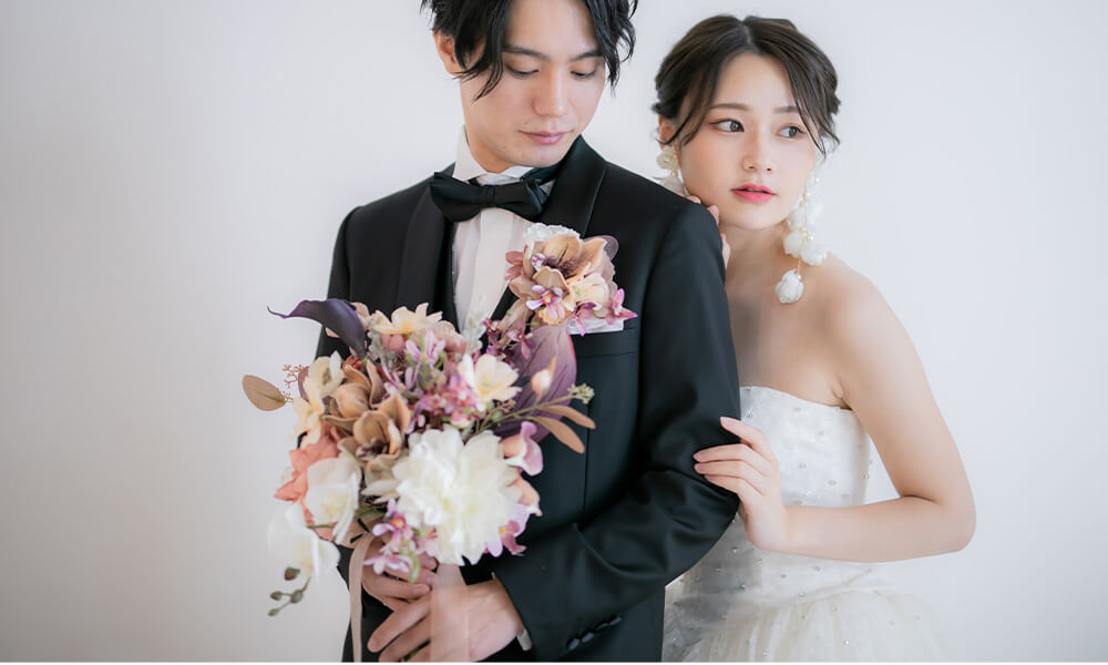 白の背景の「マイブライド岡崎光ヶ丘店」のスタジオで花束を持って洋装フォトを撮影する花嫁と新郎のイメージ