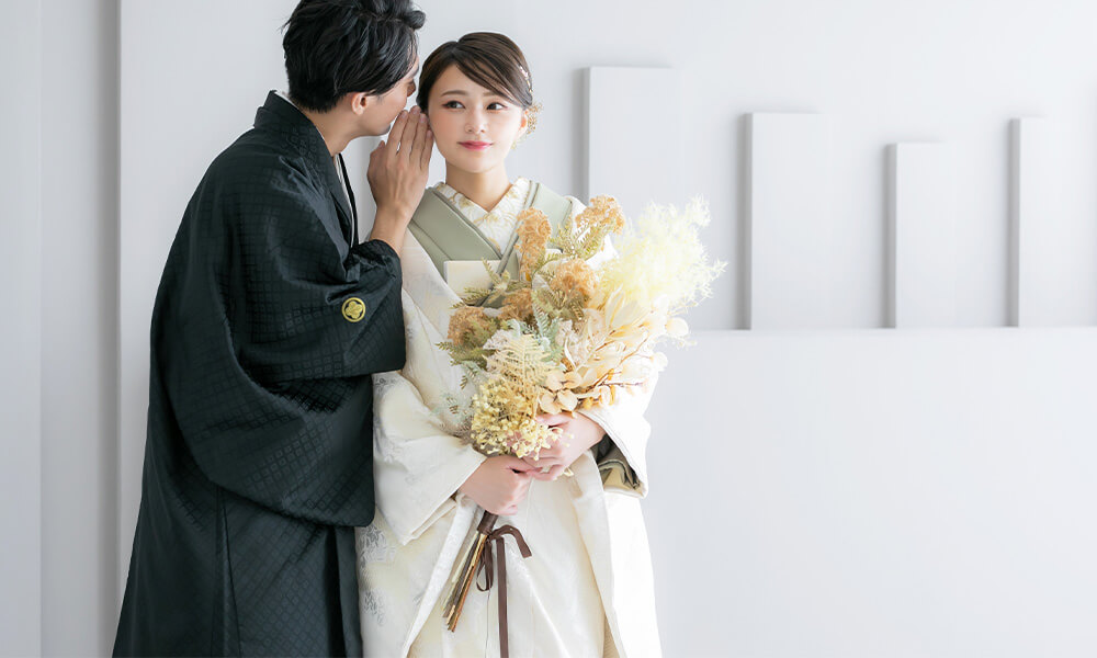 白の背景の「マイブライド岡崎光ヶ丘店」のスタジオで新郎が花嫁にささやくポーズの和装フォトのイメージ