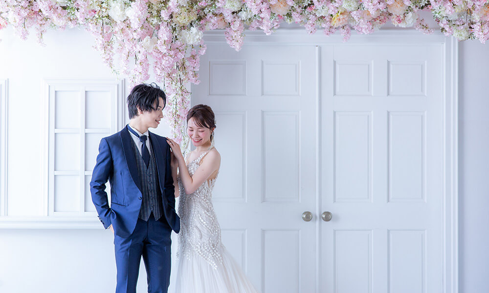 白い扉や窓を背景に、天井から下がる淡いピンクや白の花々で華やかな「マイブライド滝ノ水」のスタジオで洋装フォトを撮影する花嫁と新郎のイメージ