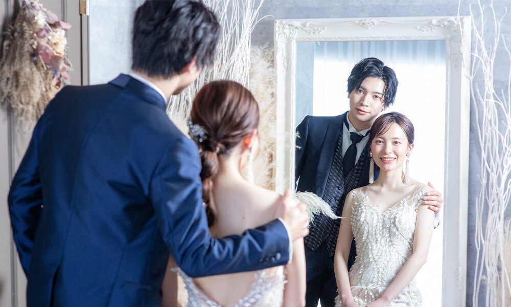 グレーの壁にシックな雰囲気の「マイブライド滝ノ水」のスタジオで姿見鏡に映り洋装フォトを撮影する花嫁と新郎のイメージ