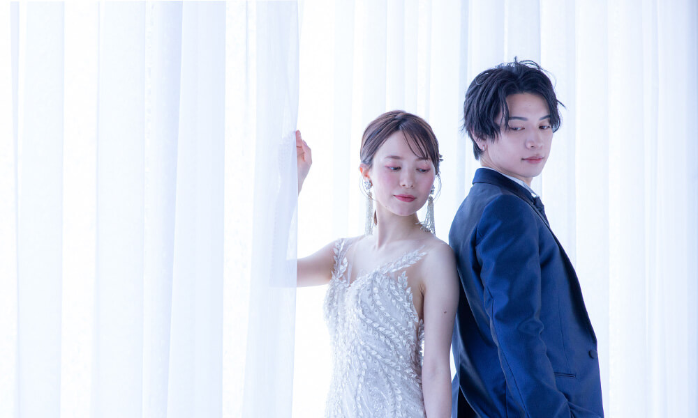 白のレースカーテンのある「マイブライド滝ノ水」のスタジオで背中合わせの洋装フォトを撮影する花嫁と新郎のイメージ