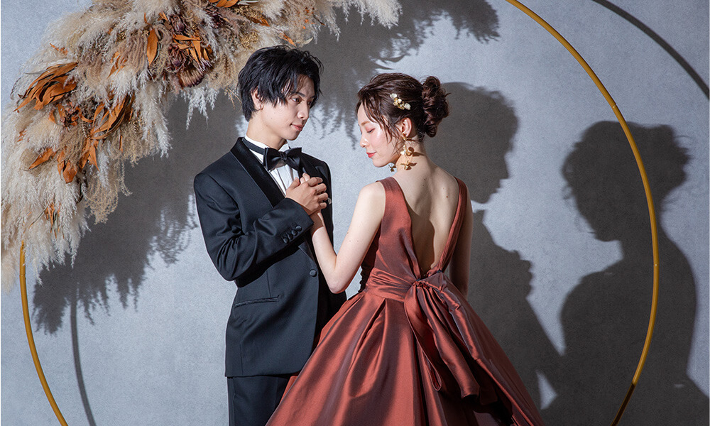 グレーの壁にドライフラワーが配置された「マイブライド天白」のスタジオで洋装フォトを撮影する花嫁と新郎のイメージ