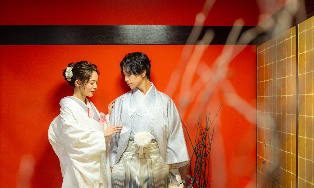 赤の背景の「マイブライド天白」のスタジオで和装フォトを撮影する花嫁と新郎のイメージ