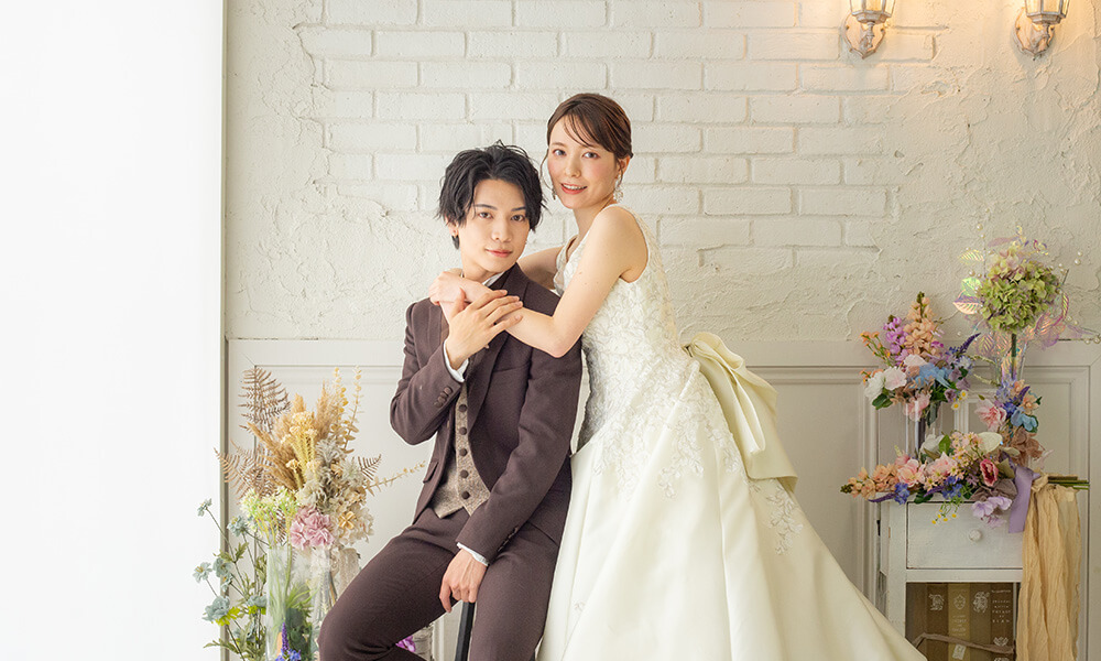 ドライフラワーが配置された白レンガの壁が美しい「マイブライド豊明」のスタジオで、洋装フォトを撮影する新郎新婦のイメージ