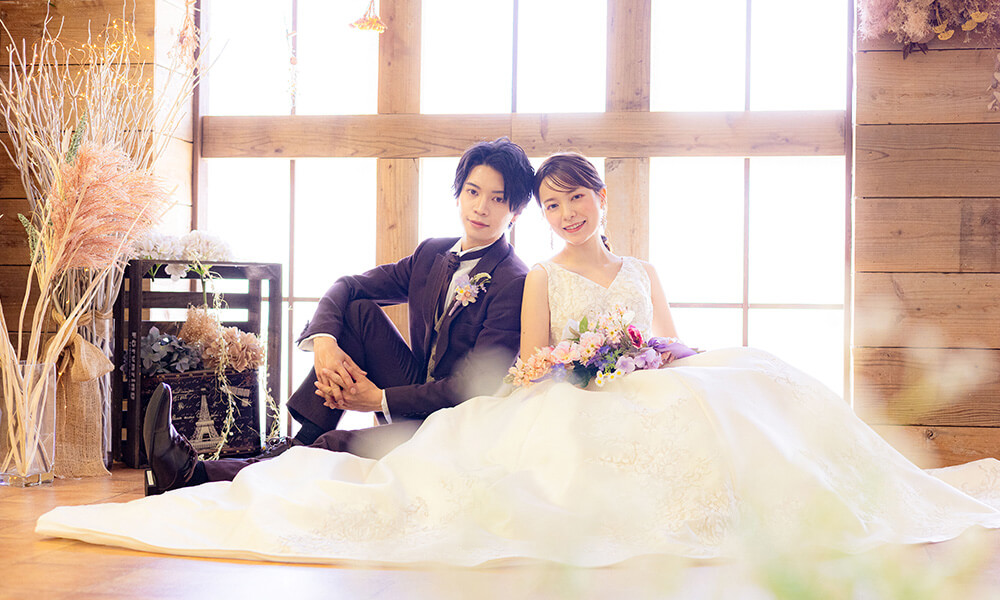ウッド調の壁にドライフラワーなどが配置された「マイブライド豊明」のスタジオで座って洋装フォトを撮影する花嫁と新郎のイメージ