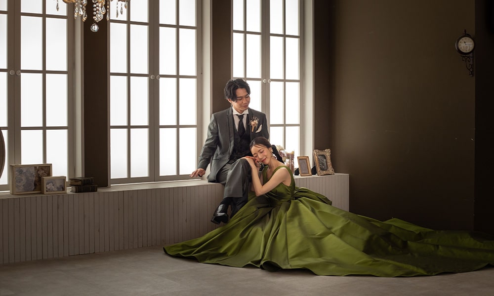 「マイブライド津」のスタジオで、窓辺に座るタキシード姿の新郎と、グリーンのドレスを着て寄り添う花嫁の洋装フォトイメージ
