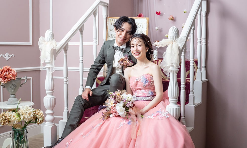 ピンクを基調とした「マイブライド津」のスタジオで、階段に座りブーケを持って寄り添う新郎新婦の洋装フォトイメージ