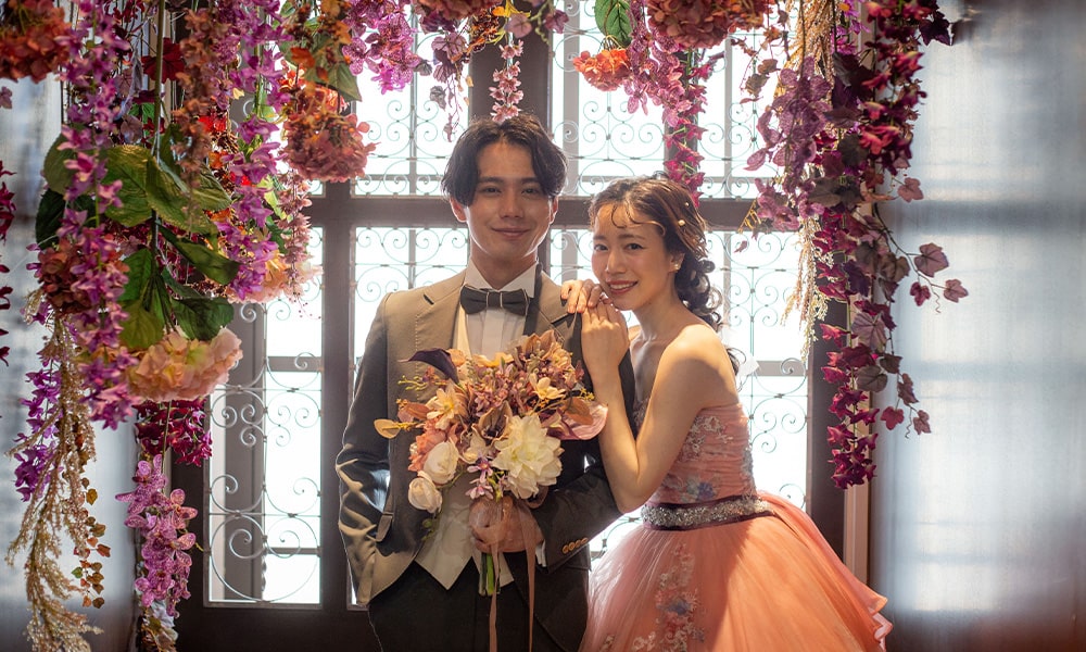 「マイブライド津」の花に囲まれたスタジオで、新郎新婦がブーケを持ち寄り添う洋装フォトイメージ